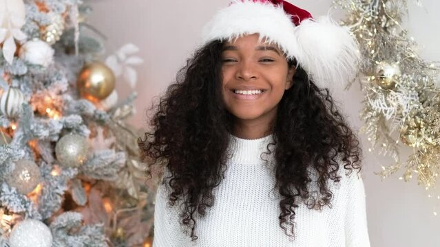 Christmas and New Year portrait beautiful smiling African American girl in Santa hat near Christmas tree, Smiling young black woman with curly hair looking at camera.