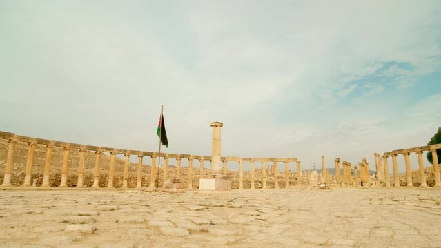 Panoramic view of ancient columns placed in Oval Plaza, Jerash, Jordan. Vast open place surrounded by pillars in antique ionic style, with flag in middle of square. 4k zoom in shot