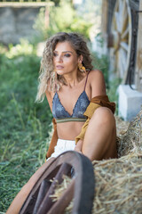 Fototapeta na wymiar Sensual girl with long legs and brown boots sit on the wood chair at farm . Handsome girl wearing beautiful body in outdoor western scene. Fashion blonde model with long legs, bra and curly hair