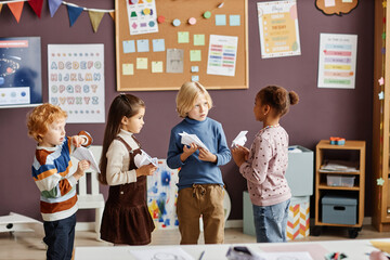 Group of four intercultural little learners of kindergarten in casualwear playing paper planes and interacting at break in classroom