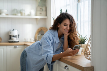 Fototapeta na wymiar Happy pleasant young woman using smartphone, enjoying online shopping, spending leisure time at cozy home. Smiling female standing in kitchen holding mobile, surfing internet or chatting with friends