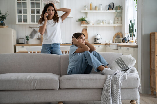 Angry strict woman mother holding smartphone making phone call to husband to discuss teenage daughter behavior, upset teen girl sitting on sofa covering ears not to listen mom lecturing her at home