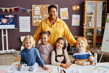 Happy mature teacher and group of intercultural little learners standing by desk and looking at...