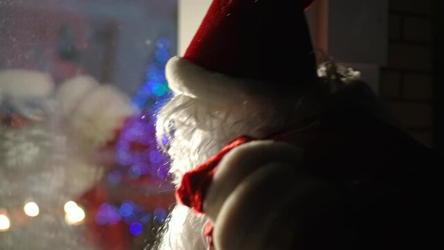 Close-up Caucasian man in Santa costume knocking in window gesturing inviting outdoors. Side view portrait of positive guy making surprise on snowy Christmas night