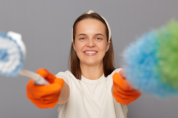 Happy female holding PP duster and brush, keeps home clean, wears orange rubber gloves, standing...