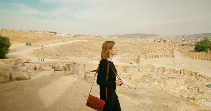 Black dressed woman visits and snaps photos of landscape and Oval Plaza in Gerasa, Jerash, Jordan. Young Tourist lady takes pictures by analogue camera of ancient ruins in summer. 4k tracking shot