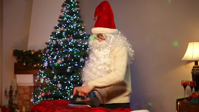 Cheerful young man dancing in living room with Christmas tree ironing Santa costume. Portrait of joyful guy having fun preparing New Year surprise at home indoors