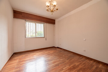 Empty living room with reddish parquet with matching skirting boards and wooden doors with aluminum barred windows and multi-arm ceiling lamp