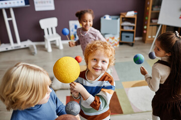 Cute smiling primary school learner with model of sun looking at camera while standing among other kids playing with planets in kindergarten