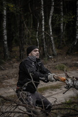 Obraz premium A man in work gloves pulls a tree branch with both hands. A Caucasian bearded man sorts out fallen trees to cut them into firewood. Men's work concept.
