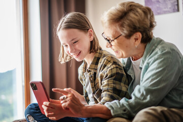 Smiling girl granddaughter with happy grandmother having fun and using looking at smartphone screen