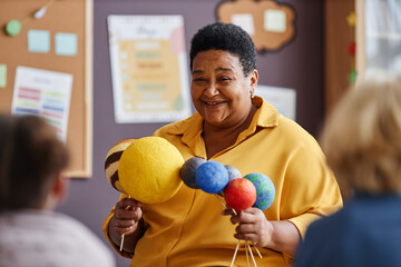 Happy mature African American teacher with planet models speaking about solar system to little...