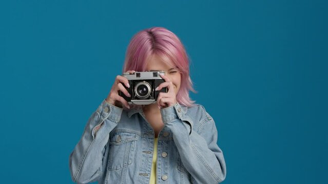 Portrait of cheerful beautiful student with analogue camera in studio. Happy pretty young hipster woman tourist taking photo. Photographer taking pictures.Female with Pink Color Hairstyle in good mood