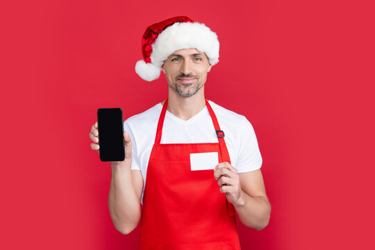 mature man in christmas santa hat and red apron hold smartphone and card. online banking