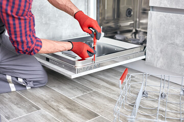Male worker in special clothes installs or removes the dishwasher. Technician repairing or fix door of dishwasher. Service man setting up dishwasher in modern kitchen.