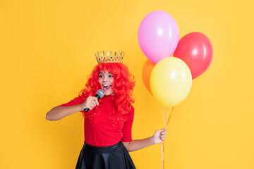 happy girl in crown with microphone and party balloon on yellow background