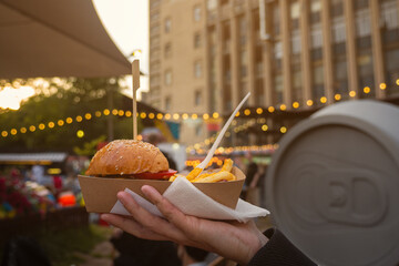 Man hands holding street food burger with french fries on craft paper. Street fast food. festival background