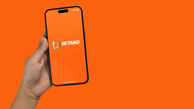 Boy holding a smartphone iPhone 14 Pro with Betano betting provider app on the screen. Orange background. Rio de Janeiro, RJ, Brazil. November 2022
