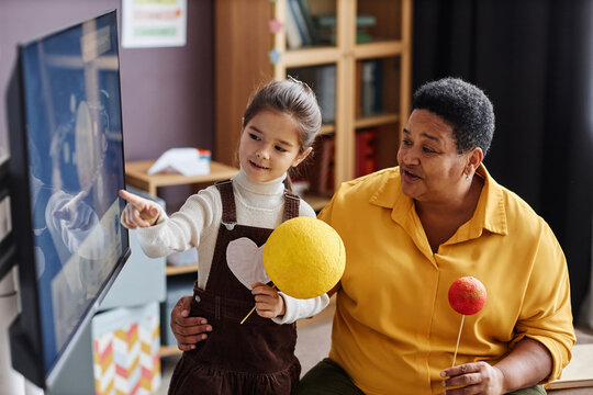 Clever little girl with model of Sun pointing at interactive board during presentation of solar system and discussion with teacher