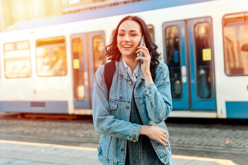 a young woman in a denim jacket is talking on the phone and waiting for a tram at the stop Lifestyle photo - obrazy, fototapety, plakaty