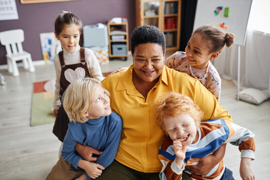 Happy mature African American teacher of nursery school embracing two adorable boys while having fun with group of intercultural kids
