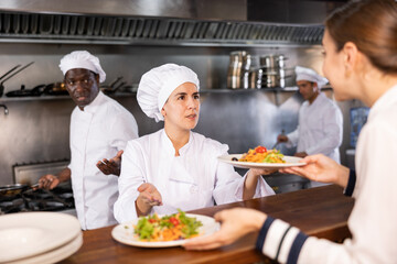 Portrait of smiling young woman chef working in restaurant kitchen, giving out ready salads s to waitress on order station