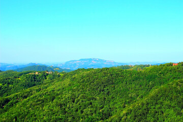 Fototapeta na wymiar Spacious view in Montemonaco at the thrilling hilly Marche landscapes suffused with lush greenery and some distant perched buildings surmounted by a crest of faded mountains in the background