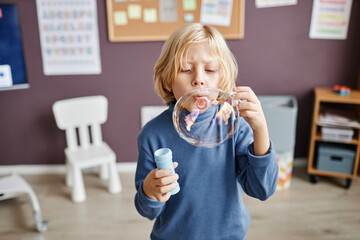 Adorable little boy with blond hair blowing soap bubbles while standing in front of camera in classroom of nursery school or kindergarten