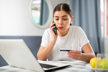 Young girl sitting at a table in front of a laptop is talking to someone on a mobile phone, dictating credit card details