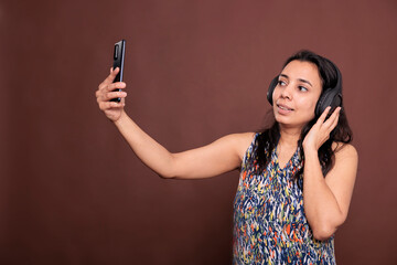 Indian woman in headphones talking on smartphone online meeting, chatting on teleconference, looking at mobile phone camera. Videocall conversation, remote communication, videoconference