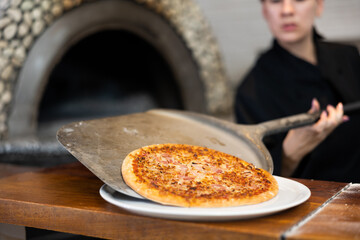 Woman chef preparing pizza in restaurant, putting freshly oven baked pizza from paddle to plate