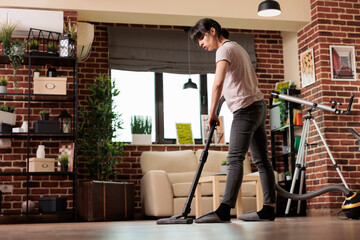 Woman using vacuum cleaner on tile floor, concentrating on spring cleaning. Housewife doing weekly cleaning routine at home, dedicated to keeping everything tidy and clean, low angle view. - Powered by Adobe