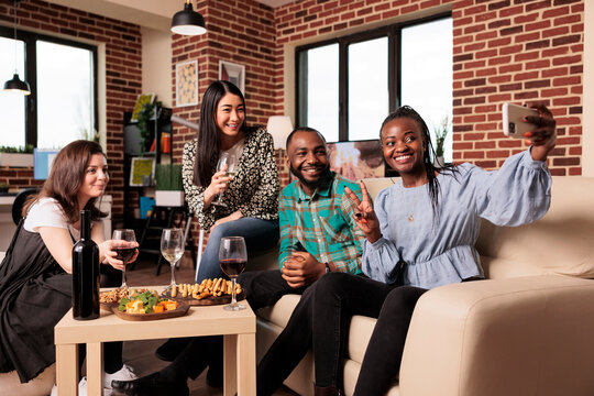 African american adult woman taking cell phone picture with asian, caucasian friends at apartment gathering. Multiple nationalities people posing for photo, drinking wine, eating snacks.