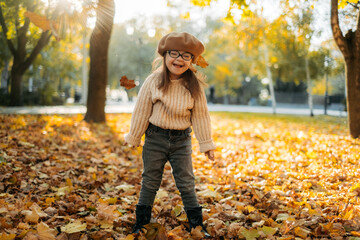 Happy cute brown-haired woman with Down's syndrome in a knitted sweater and stylish glasses throws bright foliage into the sky and laughs, the child enjoys time in a warm autumn park, happy childhood