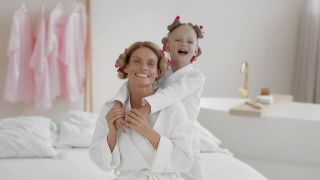 Carefree little girl embracing her attractive mother and smiling to camera, both wearing bathrobes with curlers on hair