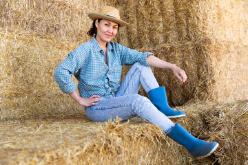 Naklejka na ściany i meble Portrait of smiling young woman farmer posing at hay storage on farm