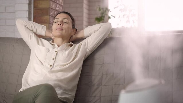 Working aromatherapy diffuser by female. A happy woman relax on the sofa by the working aromatherapy diffuser in the room.