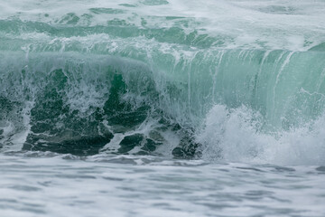 An angry turquoise green color massive rip curl of a wave as it rolls along the ocean. The white mist and froth from the wave are foamy and fluffy. The Atlantic Ocean in the background is a deep blue