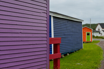 Multiple colorful wooden beach huts on vibrant green grass. The small buildings are purple, blue, orange, and white colored. The orange shed has a vibrant single green door with a small glass window.