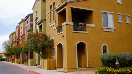A street view of a row of warm colorful townhouses with stucco coating on the outside, arched doorways, and balconies with colorful red flowering trees in front.
