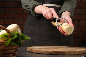 Woman peeling fresh ripe parsnip at black table, closeup