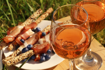 Wooden board with glasses of delicious rose wine and food on green grass outdoors, closeup
