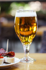 Amber beer in glass. Elegant tableware with golden freshly brewed wheat beer stands on wooden table in cafe, bar pub terrace. Alcoholic beverage with traditional Spanish dish tapas, sausage sandwiches