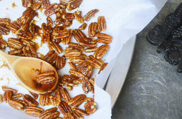 Fresh candied pecans on parchment paper covered in decadent brown sugar sauce. A wooden spoon has some nuts. Background is a vintage zinc countertop. Antique cast iron trivet in corner