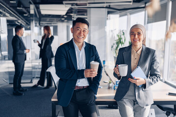 Two happy multiracial colleagues talking and drinking coffee during break time in office