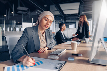 Focused muslim businesswoman in hijab working with documents while sitting in modern office