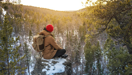 Fototapeta premium A tourist-traveler with a backpack in a red hat sits on the edge of a cliff and looks at a forest winter valley