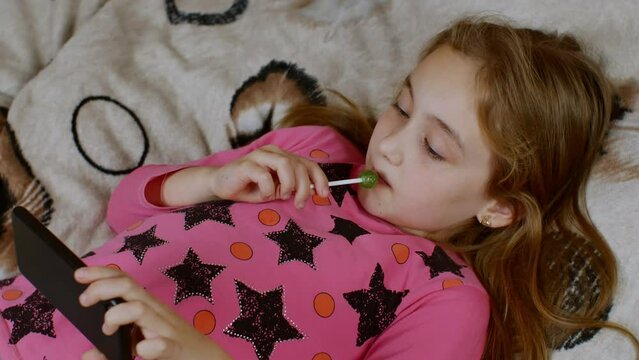 A cute girl with long hair is lying on the sofa, carefully looking at the smartphone screen and sucking on a sweet lollipop. The concept of digital addiction in children and adolescents