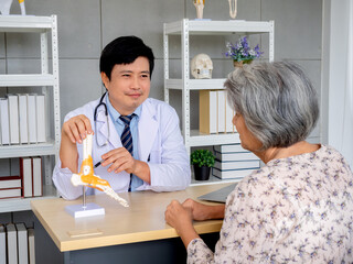 Smiling professional Asian man orthopedic doctor pointing to foot skeleton, ankle joint anatomy model to explain for senior female patient in medical office. Physio treatment, health care concept.