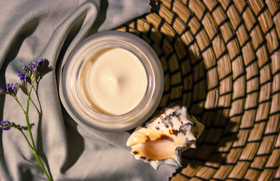 Cosmetic composition. Face cream jar, moisturizing, nourishing body cream on rattan background top view decorated with cray silver fabric, flower, seashell. Natural spa treatment. Organic cosmetics.